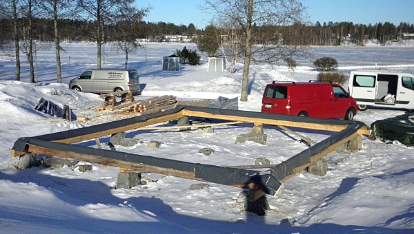 Talvinen kuva työmaalta. Kolme pakettiautoa ja ensimmäinen varvi hirsiä on pystytetty kivien päälle.