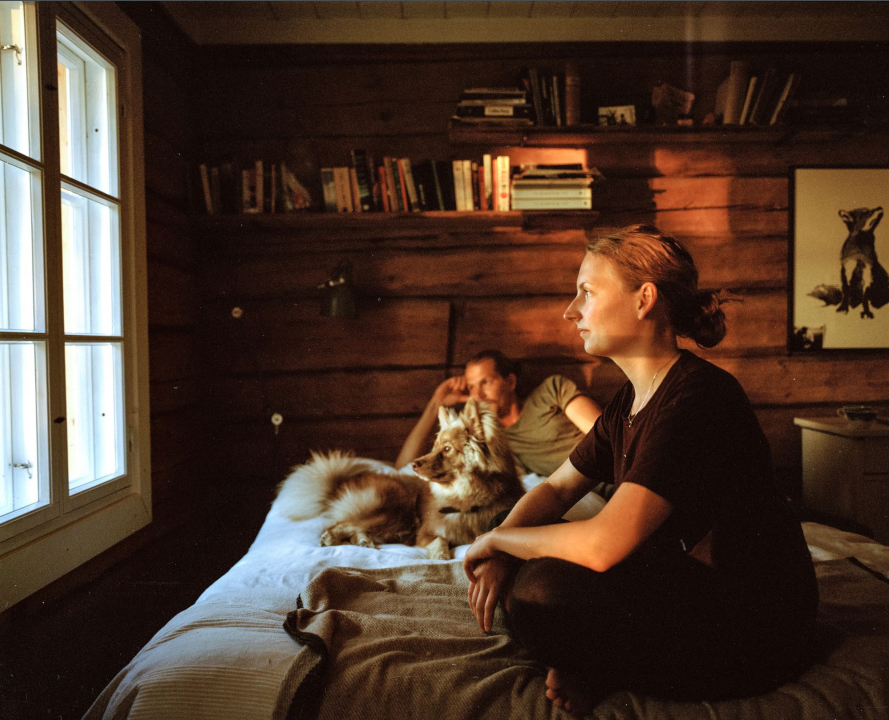 puutalossa olevalla sängyllä istuu nainen ja makoilee mies ja koira. Kaikki kolme vaikuttavat katselevan ulos viereisestä ikkunasta. Seinällä on maalaus ketusta sekä pari hyllyä, jotka ovat täynnä kirjoja.
