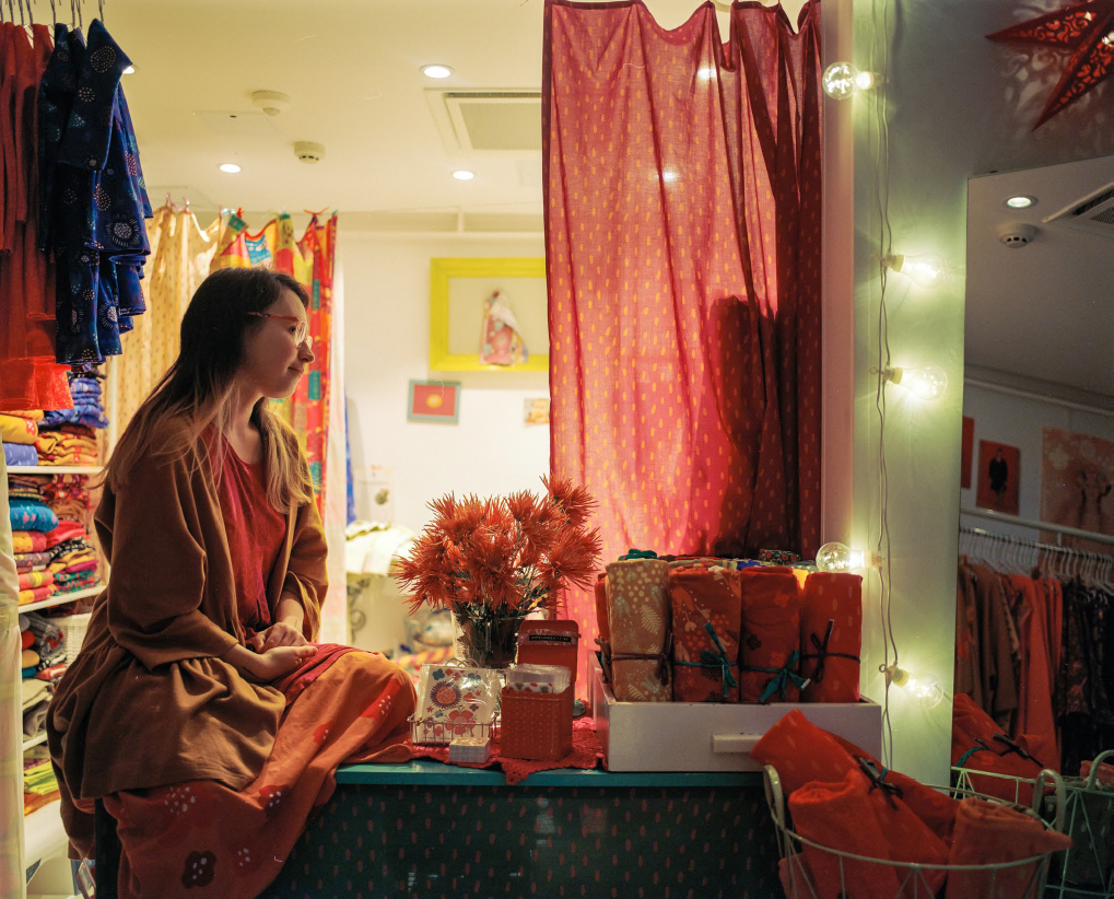 Punaisiin ja oranssiin pukeutunut nainen istuu huoneessa, jossa on suuri määrä kankaita hyllyissä ja vaatteita henkareissa.