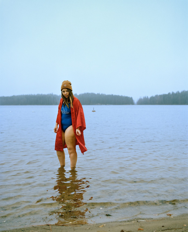 Uimapukuun, takkiin, ja pipoon pukeutunut nainen seisoo nilkkoihinsa asti vedessä. Kuva otettu rannalta ja kaukana vastarannalla näkyy metsää.