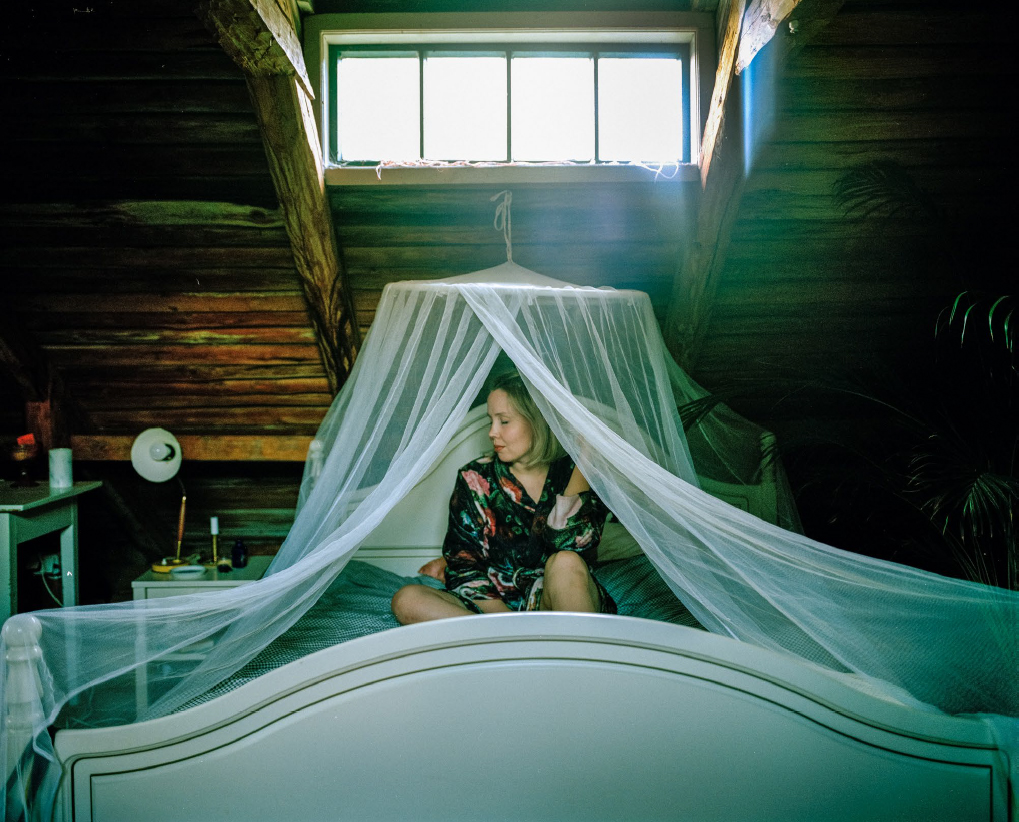 Nainen istuu puisessa, hunnulla verhotussa sängyssä ikkunan alla. Kuva näyttää olevan puutalon vintiltä tai muuten korkeimmasta kerroksesta.
