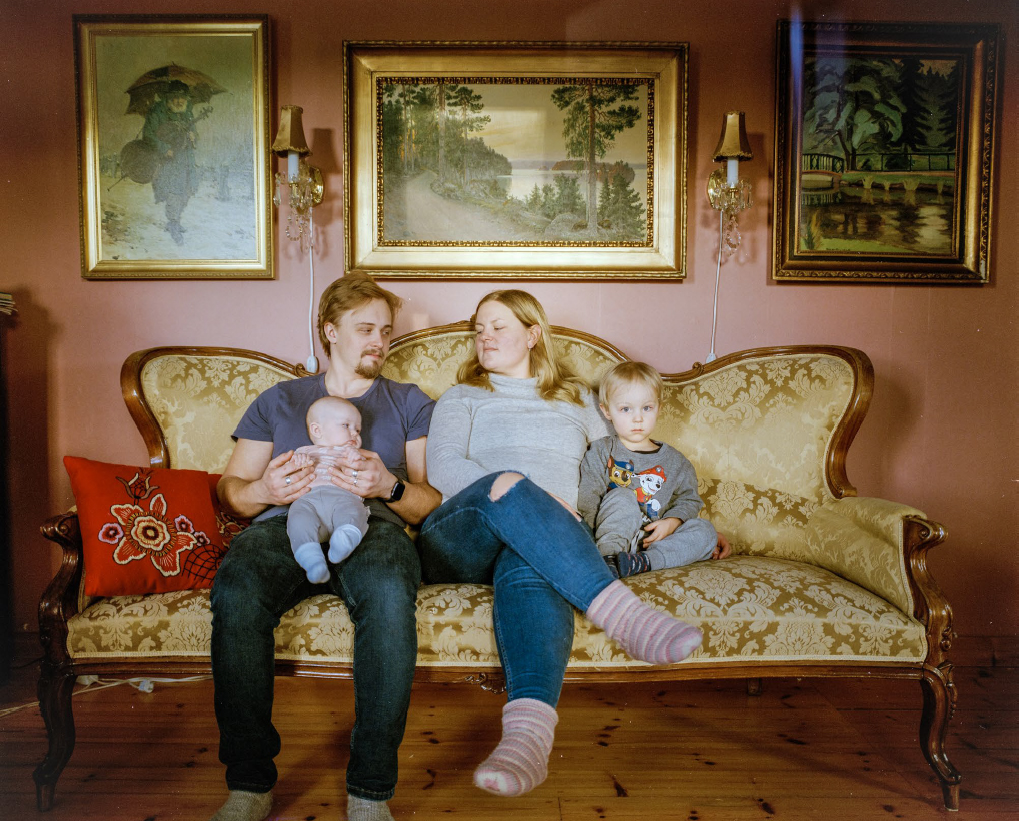 Oletettu pariskunta koostuen miehestä ja naisesta istuu kahden pienen lapsen kanssa vanhan näköisellä sohvalla. Nuorempi lapsi on vielä vauva ja mies pitää häntä sylissään. Sohvan takana olevalla seinällä on kolme maalausta ja kaksi lamppua.