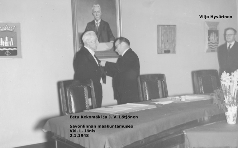 Mustavalkovalokuva, jossa kaupunginvaltuuston uusi puheenjohtaja J. V. Lötjönen kiinnittää jotain väistyvän puheenjohtajan Eetu Kekomäen rintapieleen – Viljo Hyvärinen seuraa sivusta.