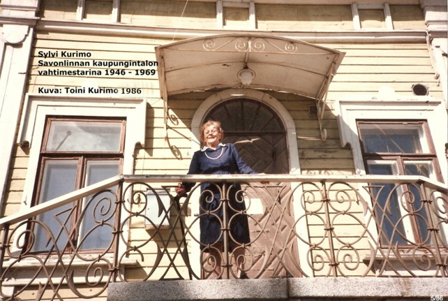 Värivalokuvassa kaupungintalossa vahtimestarina vuosina 1946–1969 työskennellyt Sylvi Kurimo pääsisäänkäynnin portaikon ylätasanteella metallikaiteen ääressä.