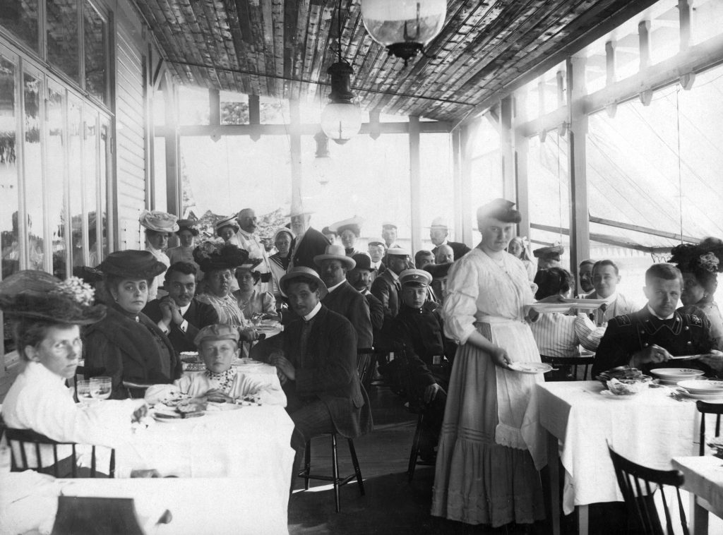 Mustavalkoinen kuva suuresta joukosta tyylikkäästi pukeutuneita ihmisiä nauttimassa ruuasta pöytien ääressä katetulla terassilla. Suurin osa heistä katsoo kameraan.