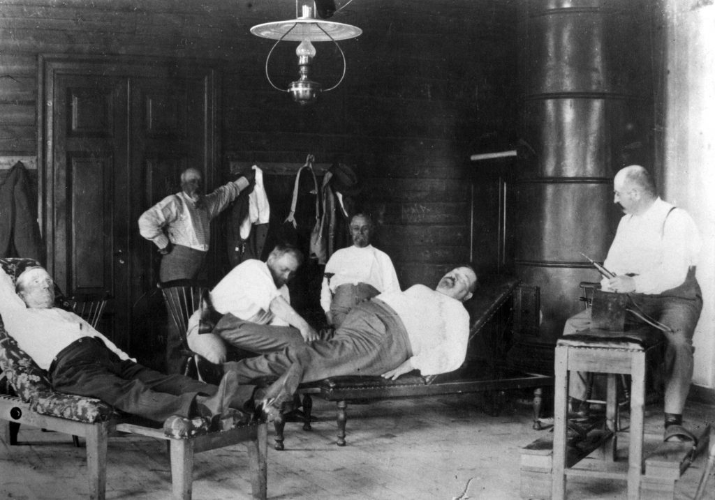 Mustavalkoinen kuva, jossa kuusi valkopaitaista miestä huoneessa. Heistä kaksi makoilee lepotuoleissa, kolme istuvat muissa tuoleissa, ja viimeinen seisoo huoneen oven lähellä.
