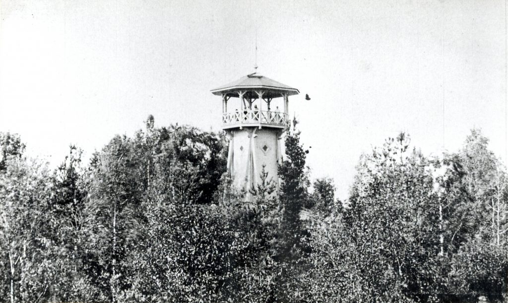 Mustavalkoisessa kuvassa näkyy puiden joukossa korkea torni. Tornin huipulla on katollinen tasanne, jolla näkyy muutamia ihmisiä.