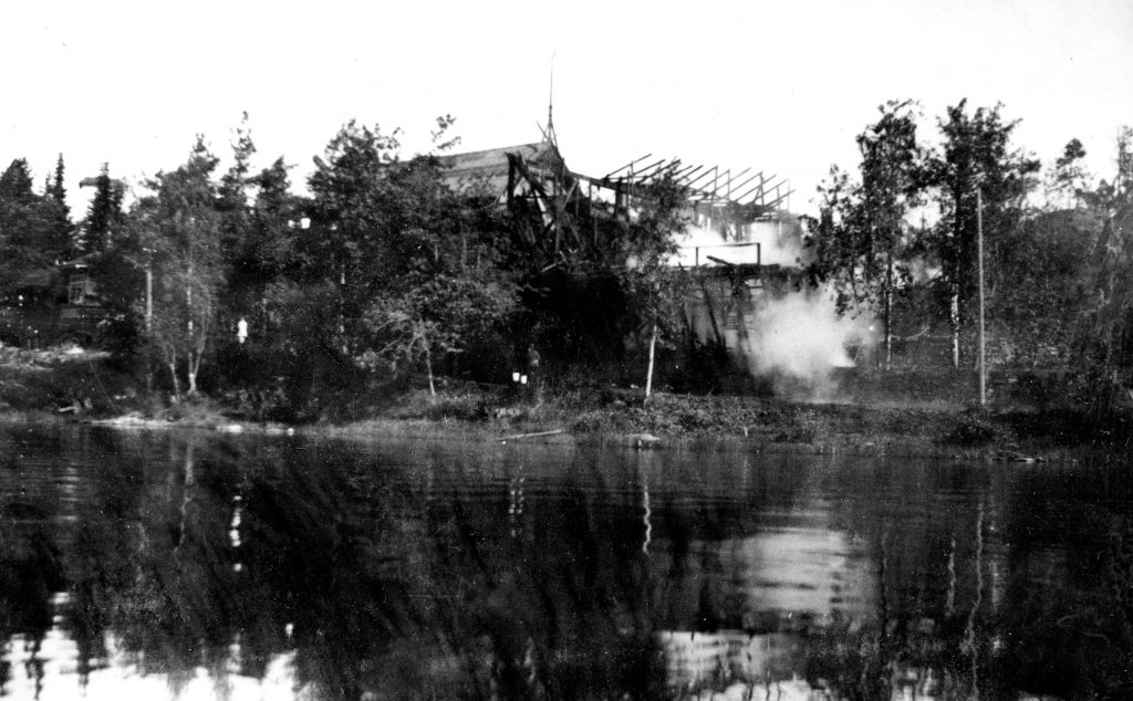 Mustavalkoisessa järveltä kuvatussa kuvassa näkyy pahasti palanut rakennus puiden keskellä. Raunioista nousee savua.