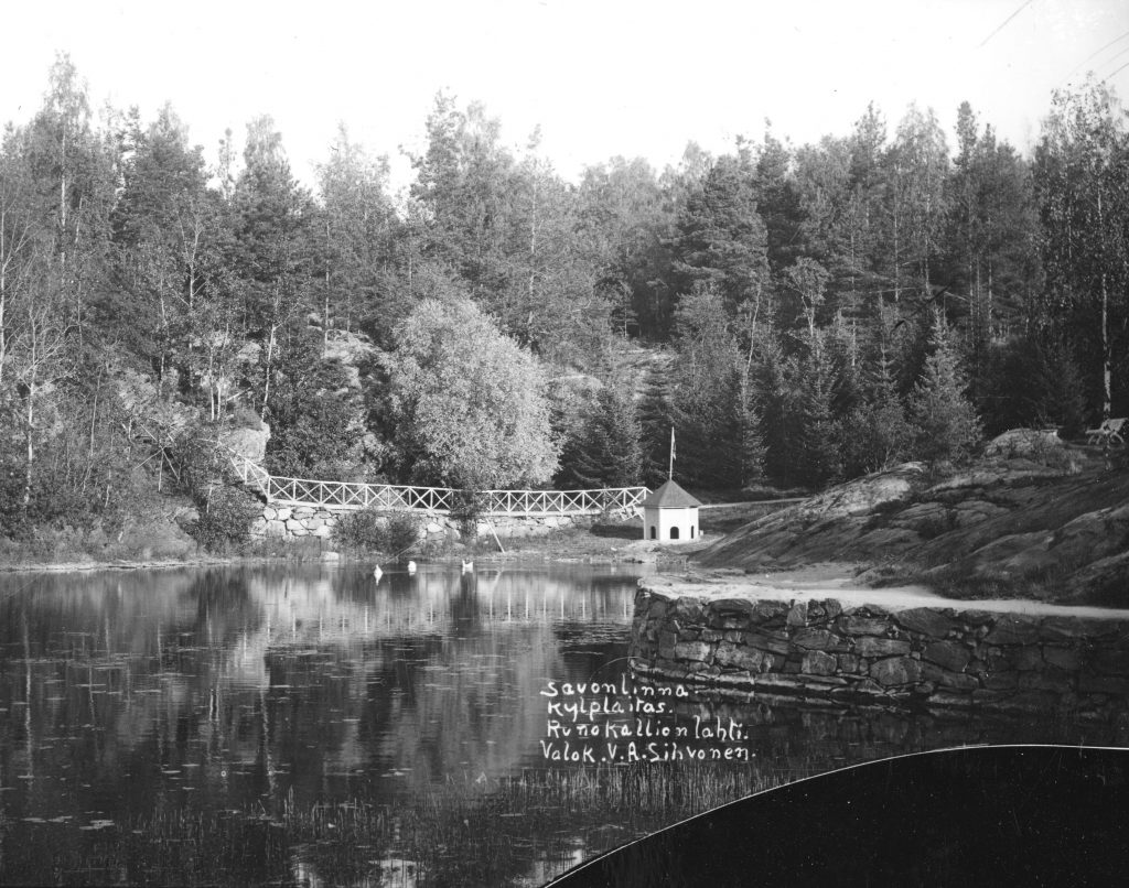 Mustavalkoisessa kuvassa on järveltä kuvattu metsämaisema. Rantaa pitkin kulkee kivien päälle tehty polku, ja kauempana näkyy silta ja pieni rakennus.