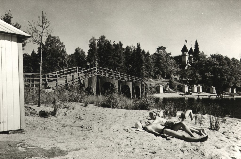 Mustavalkoisessa kuvassa näkyy ranta, jolla kolme ihmistä makailee uimavaatteissa. Taustalla näkyy silta vesialueen yli, ja sen toisella puolella uimavajoja sekä suuri puinen rakennus. Ympäristössä on myöskin reilusti puita.
