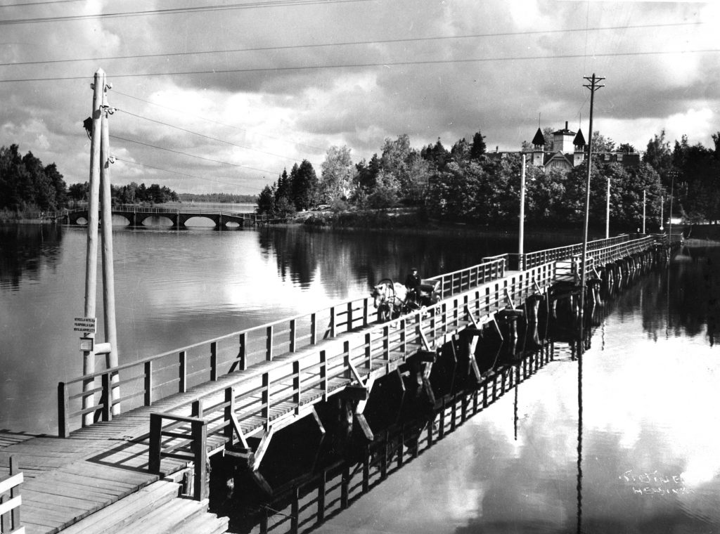 Mustavalkoisessa saaristomaisemassa näkyy kaksi puista siltaa. Lähemmällä sillalla kulkee hevosen vetämä kärry, jossa matkustaa yksi ihminen.