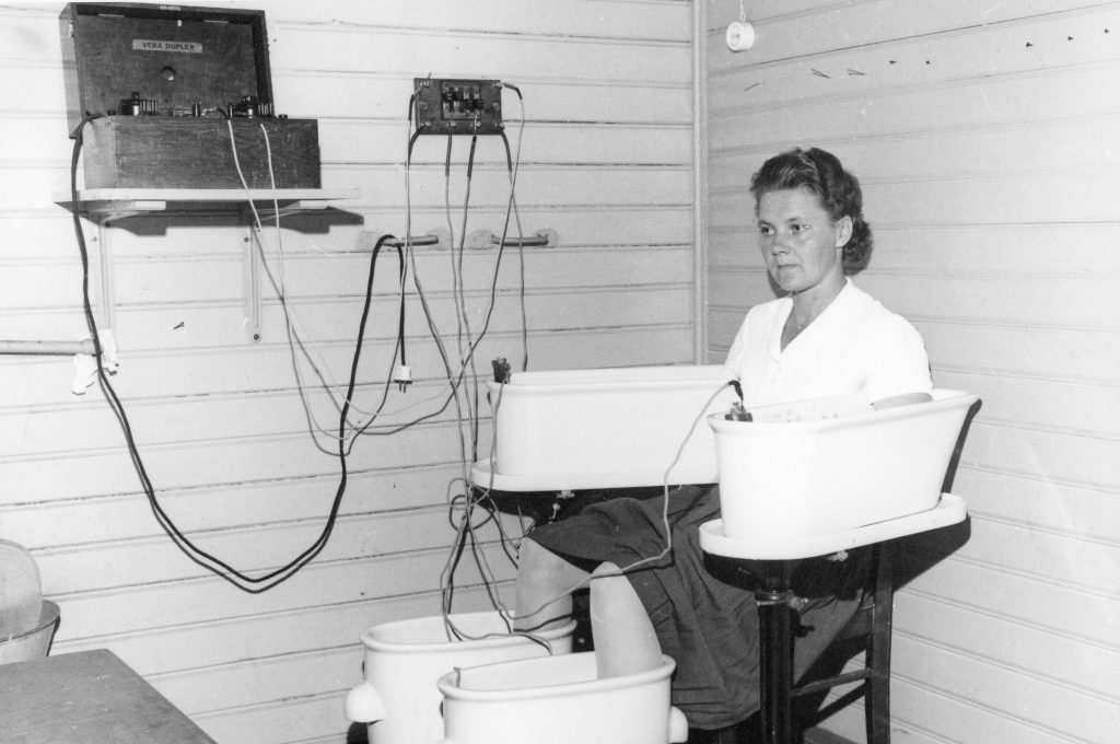 Mustavalkoinen kuva, jossa nainen istuu huoneessa tuolilla, hänen käsien ja jalkojen ollen astioissa. Astioista kulkee sähköjohtoja hyllyllä olevaan avonaiseen laatikkoon ja seinässä olevaan sähkötauluun.