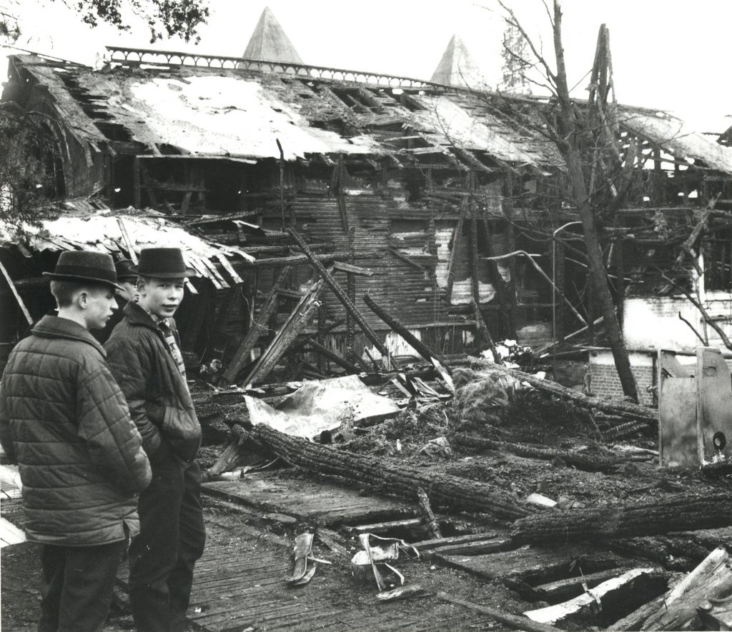 Mustavalkoisessa kuvassa näkyy kaksi miestä tutkailemassa pahasti palaneen puutalon raunioita.