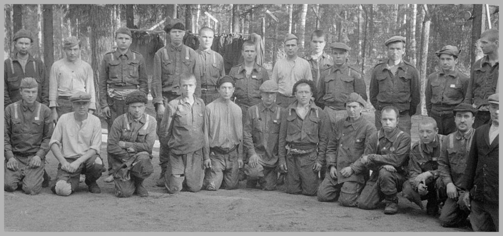 Mustavalkovalokuva venäläisistä sotavangeista, jotka ovat asettuneet kuvausta varten. Eturivissä miehet ovat polvillaan, takarivin miehet seisovat.