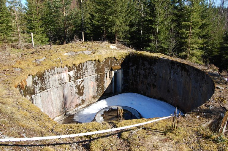 Syvä mörssäriaseman betonivalu kuusimetsän keskellä. Aseman keskellä on mörssärin jalusta ja aseman sisässä on vähän lunta.