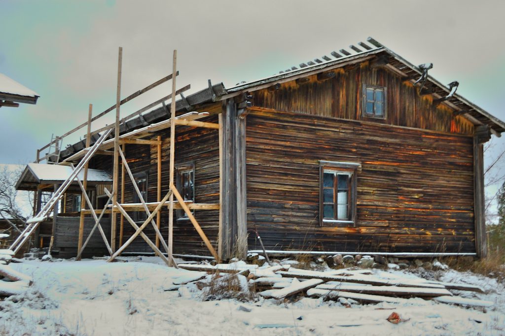 Osin lumenpeittämä hirsirakennus, jonka tuohi-malkakattoa on ryhdytty kunnostamaan. Rakennustelineet on tehty puusta.