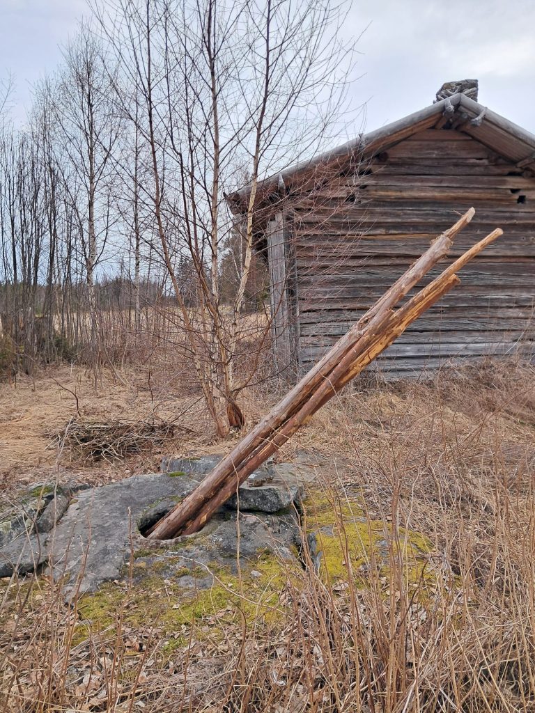 Kaivo, jonka kiven kannen aukosta on työnnetty juuriskoukkuja kaivoon. Takana hirsirakennuksen päätyä.
