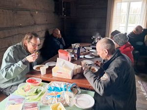 Miehiä ja naisia istuu penkeillä hirsiseinäisen tuvan pöydän ääressä syömässä.