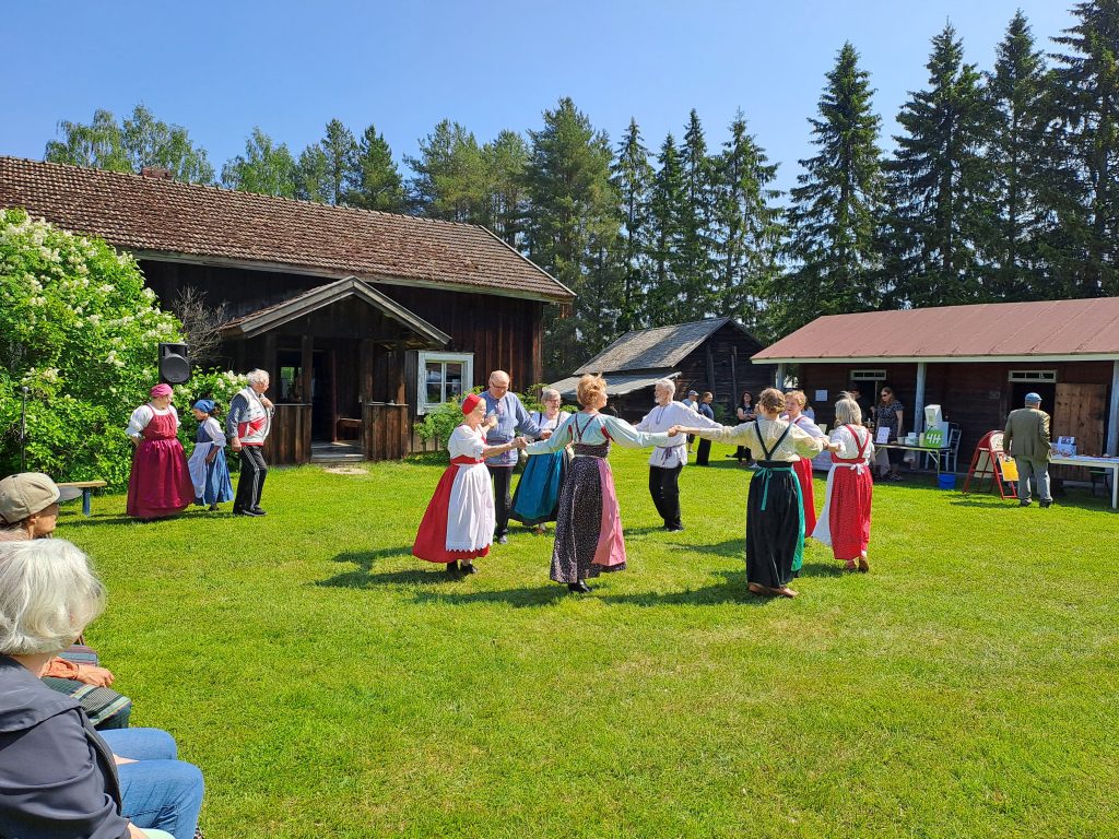 Miehet ja naiset tanssivat piirissä aurinkoisena kesäpäivänä museon nurmella – yleisö katselee.