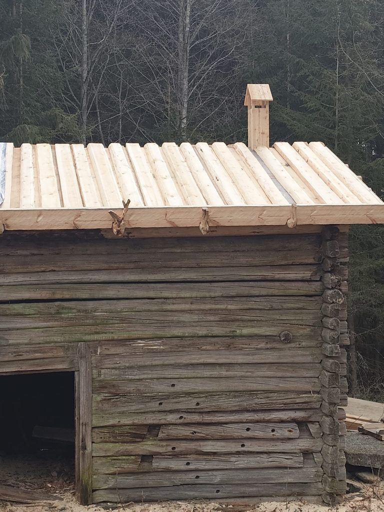 Osakuva maalaamattomasta hirsirakennuksesta, jonka toiseen päähän on uusittu lautakatto.