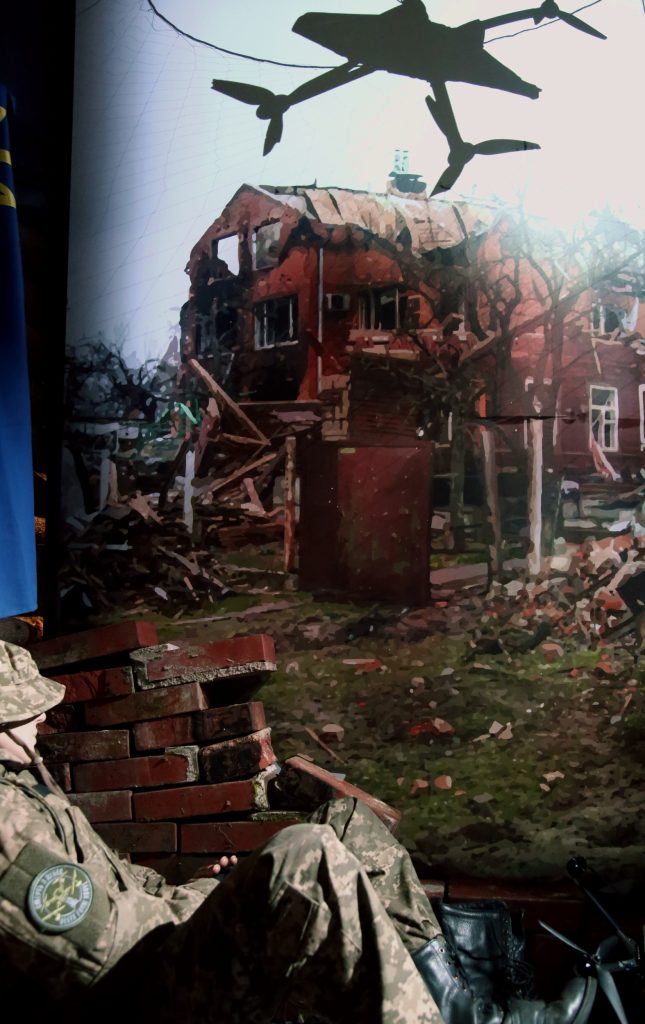 Droonin ohjaaja on suojautunut tiilimuurin taakse, ylhäällä drooni, etualalla tuhoutunut rakennus.