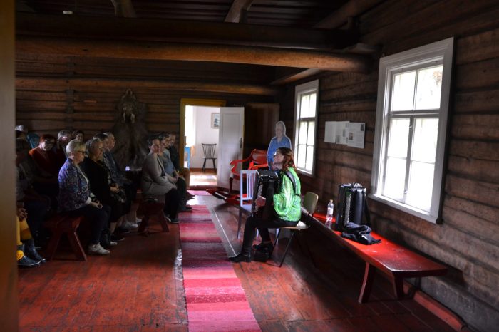 Nainen istuu harmonikan kanssa tuolilla maalaistalontuvassa ja soittaa penkeille kerääntyneelle yleisölle.