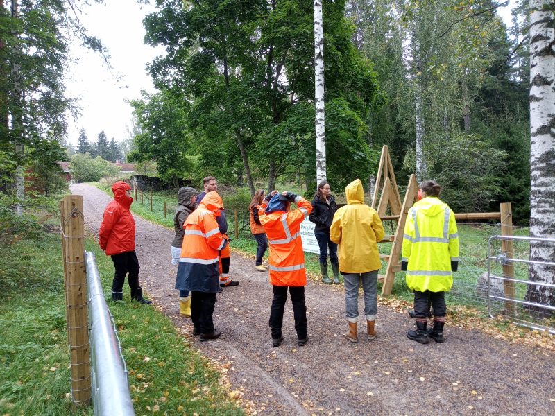 Pappilan perinnebiotoopista kertova valkopohjainen kyltti teräslankaverkkoaitaan kiinnitettynä. Taustalla vähäpuustoinen laidunalue.