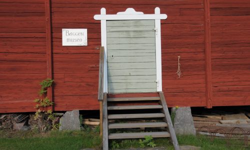 Baggen museon sisäänkäynti eli puuportaat nousevat harmaanvihreälle makasiinin ovelle, joka vuorilaudat on maalattu valkeiksi. Hirsiseinä on punamullattu, ja perustuksen ovat välikivet lohkokivistä.