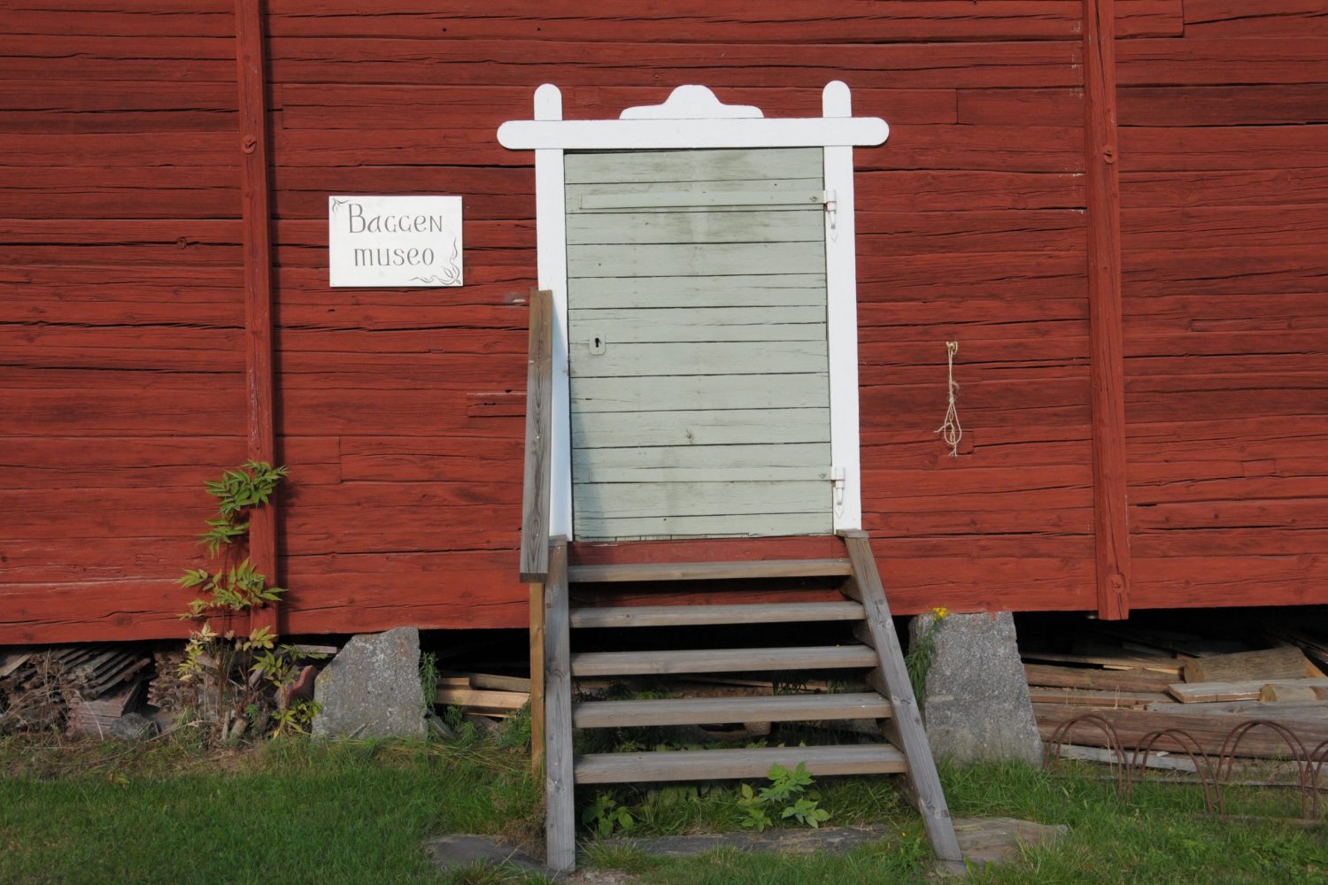 Punainen rakennuksen seinä, jossa olevalle ovelle nousee pieni puinen portaikko. Rakennus näyttäisi olevan kivien päällä ja sen alla on lautoja. Oven vieressä on kyltti, jossa lukee "Baggen museo" ja rakennuksen edessä on ruostunut pyöräteline.