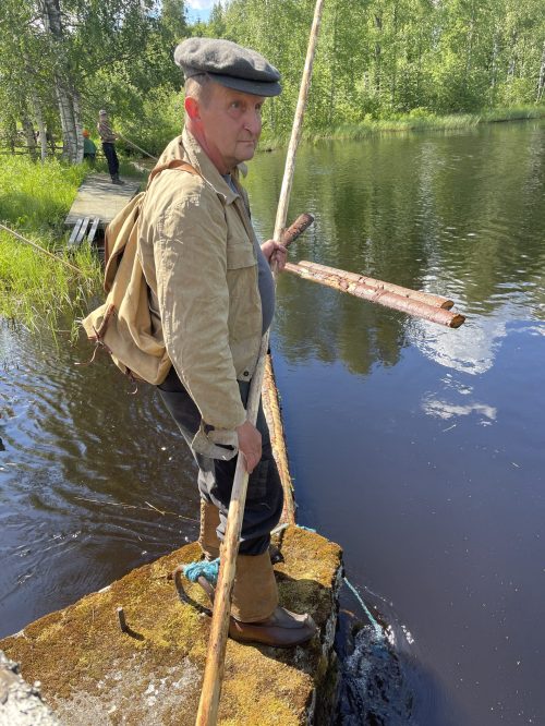 Mies reppu selässä, nahkasaappaat jalassa ja keksi kädessä veden äärellä. Taustalla tukkeja vedessä.