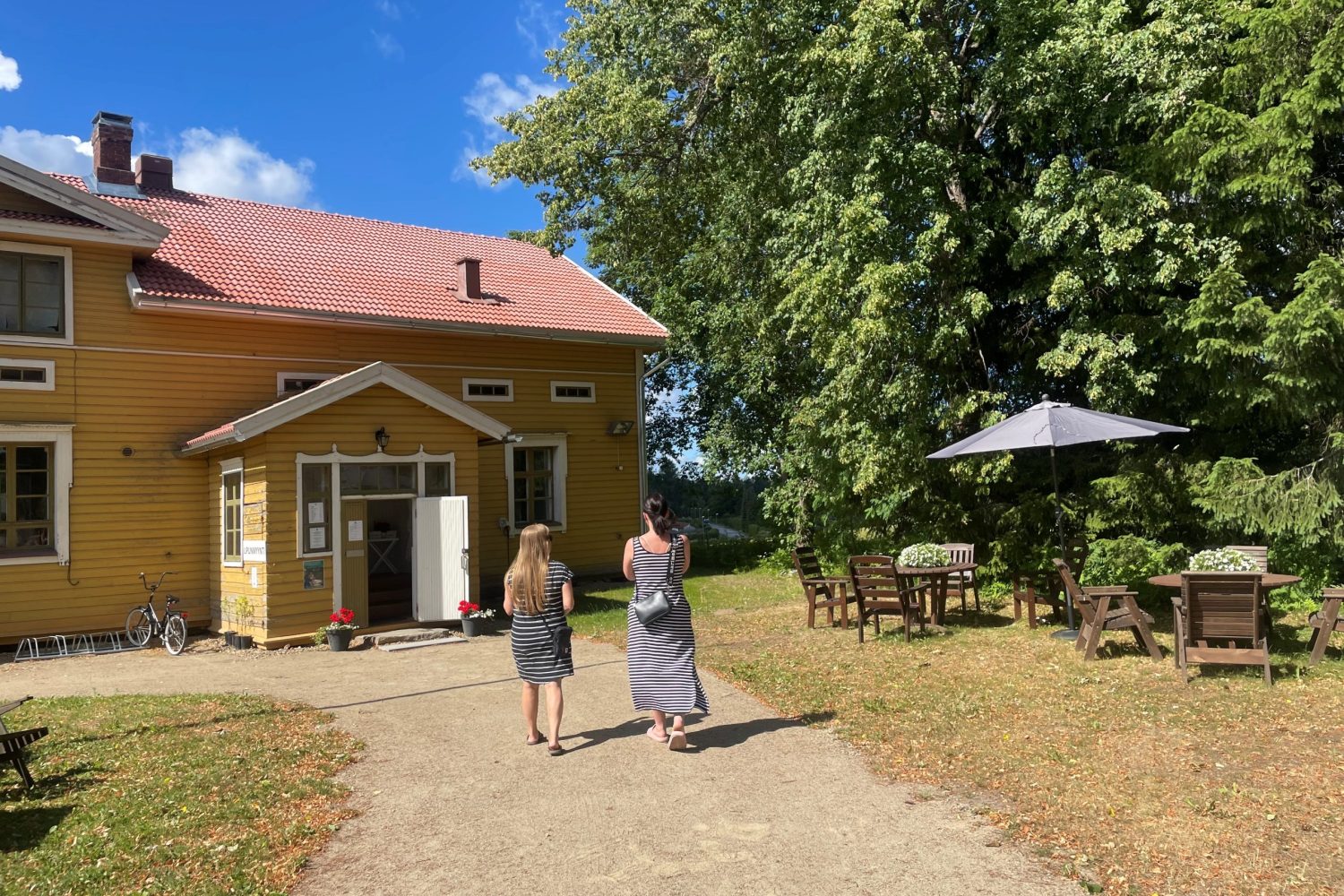 Kaksi ihmistä kävelee hiekkakäytävää kohti keltaiseksi maalattua puurakennusta. Heidän oikealla puolellaan on pöytiä tuoleja ja aurinkovarjo.