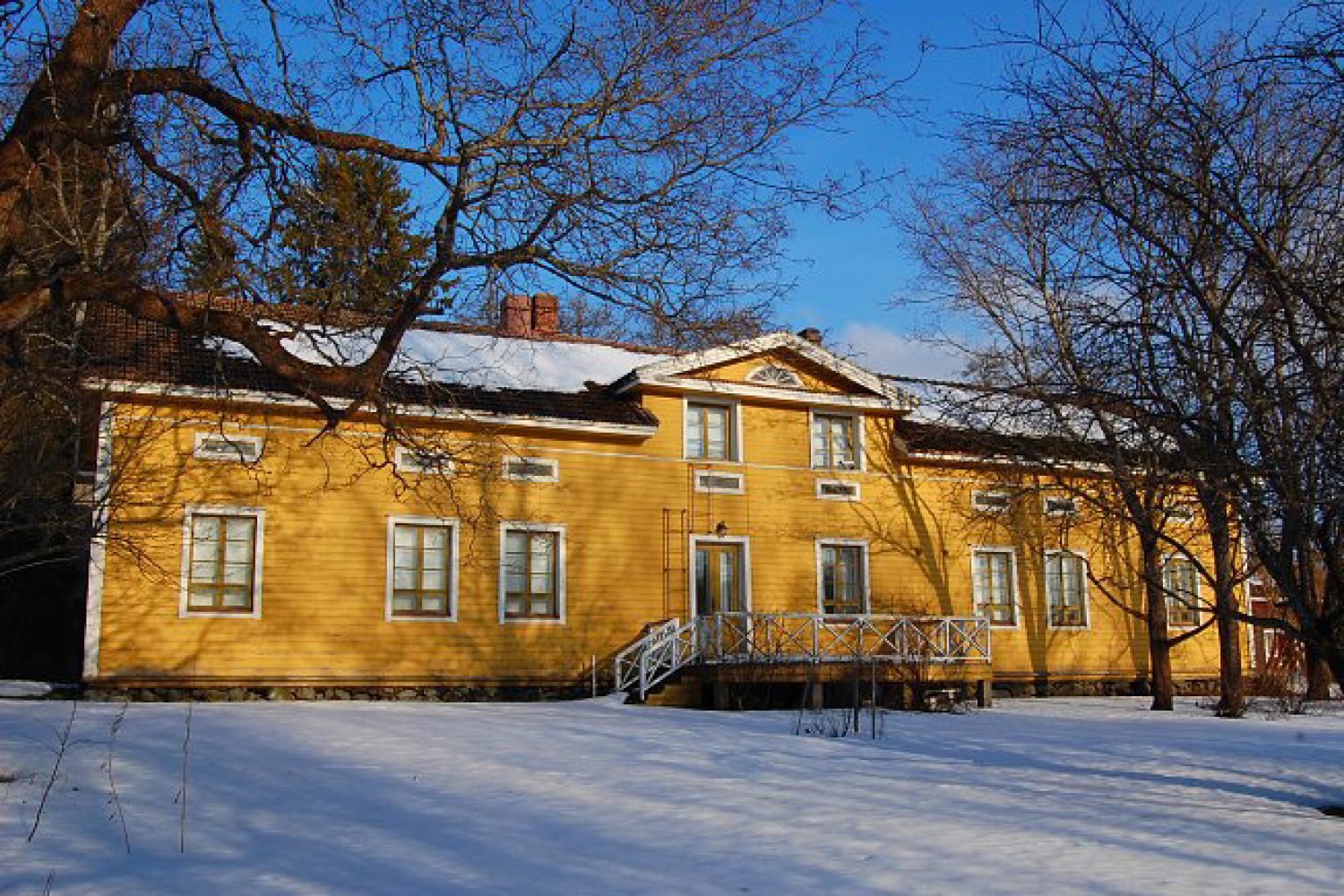 Keltainen suuri talo talvimaisemassa. Rakennuksessa on oven luona kuisti ja näkyvässä seinässä on yhdeksän ikkunaa.