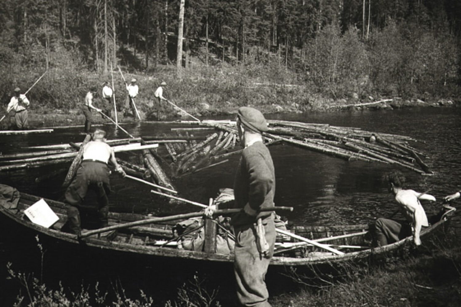 Mustavalkoisessa kuvassa joukko miehiä työskentelevät tukkien uittamisen parissa ohuella vesialueella. Miehillä on keksit ja soutuvene apunaan.