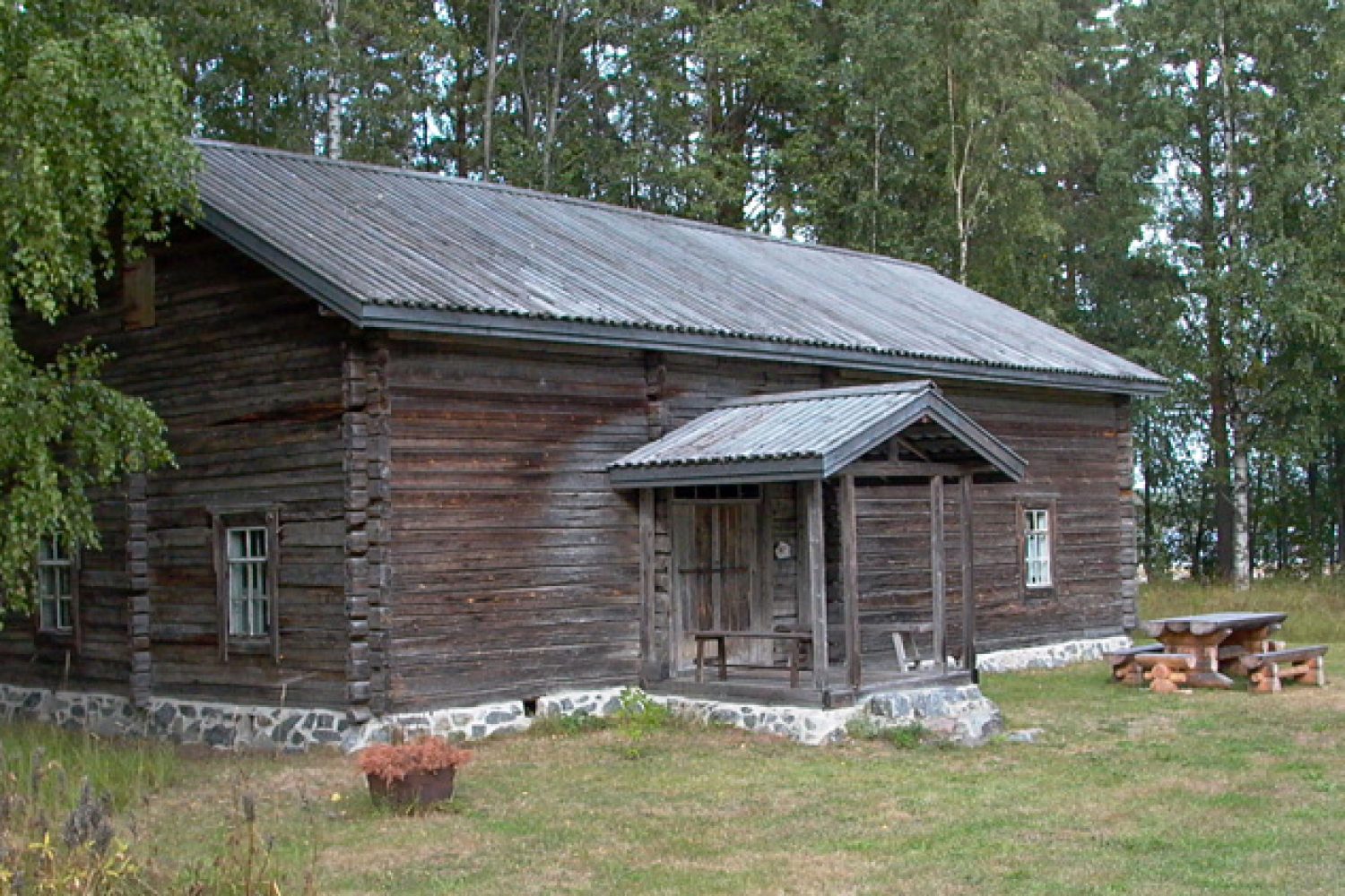 Puinen talo metsämaisemassa. Talossa on kuisti ja kolme ikkunaa. Sen edessä on puinen pöytä ja kaksi penkkiä.