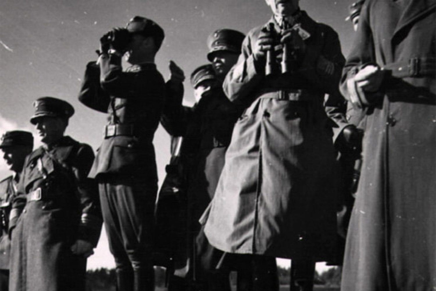 Mustavalkoisessa kuvassa kahdeksan sotapukuista miestä seisoo ulkona. Kaksi heistä pitelevät kiikareita käsissään.