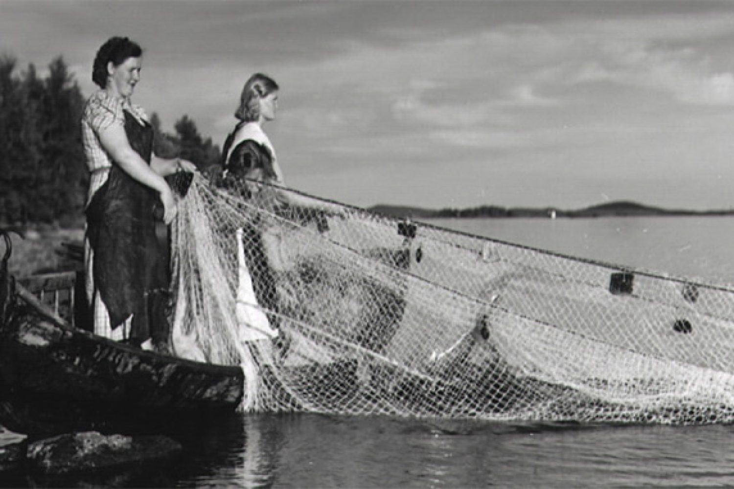 Mustavalkoisessa kuvassa kaksi naista vetävät kalaverkkoja soutuveneen kyytiin.