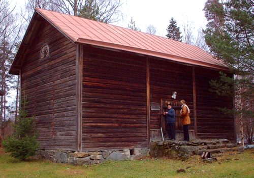 Ulkokuva maalaamattomasta ja vuoraamattomasta hirsirakennuksesta eli Enonkosken kotiseutumuseosta. Rakennuksessa on punainen saumapeltikatto, kivitä muuratut portaat.