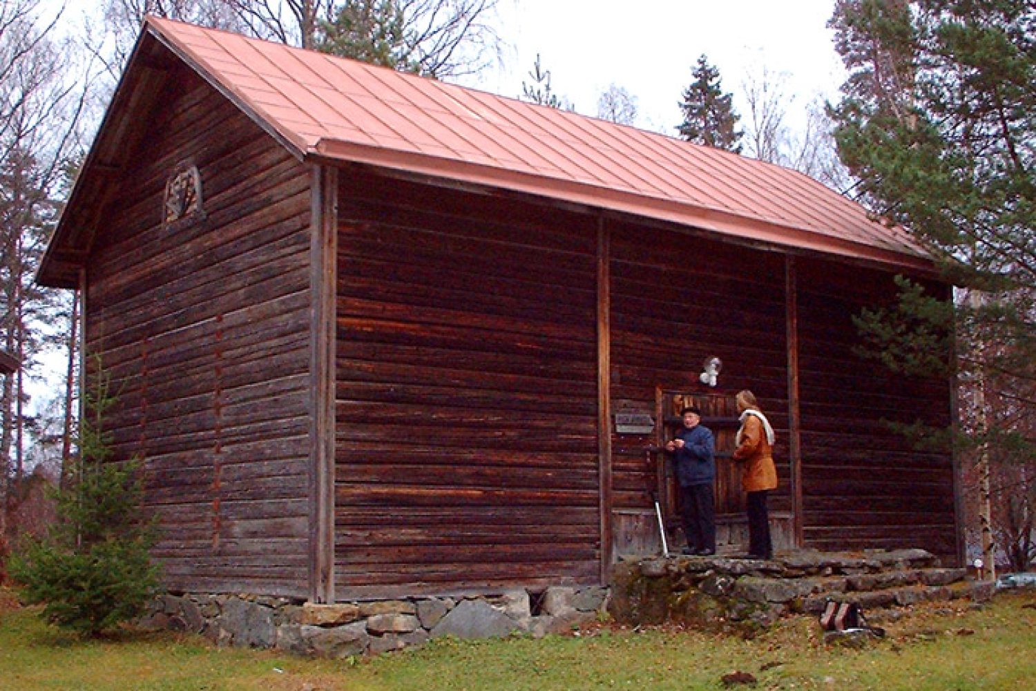 Ulkokuva maalaamattomasta ja vuoraamattomasta hirsirakennuksesta eli Enonkosken kotiseutumuseosta. Rakennuksessa on punainen saumapeltikatto, kivitä muuratut portaat.