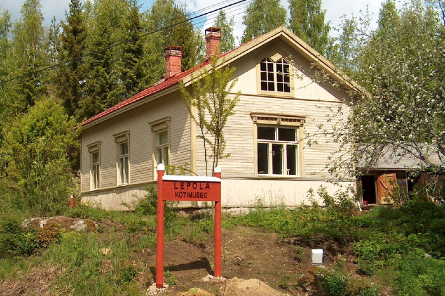 Lepolan päärakennus tieltä kuvattuna keväällä. Edessä punavalkoinen, puusta valmistettu viitta, jossa lukee Lepola kotimuseo.