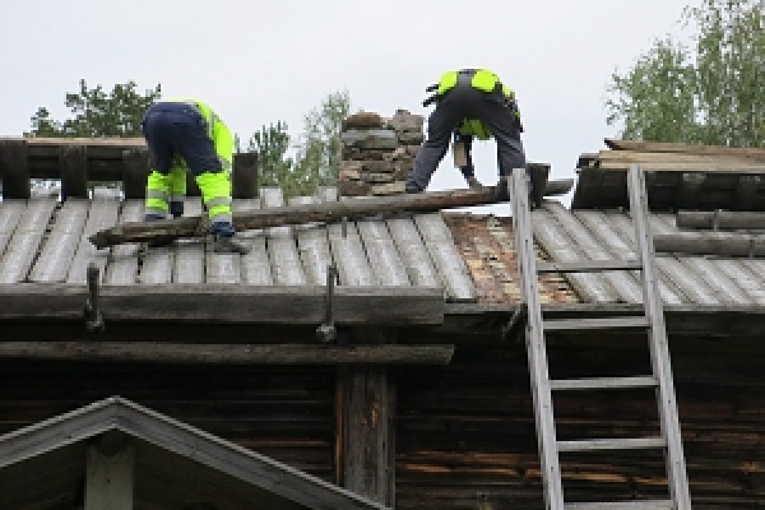 Kaksi miestä on asuinrakennuksen tuohi-malkakatolla siirtämässä painomalkoja pois paikoiltaan.