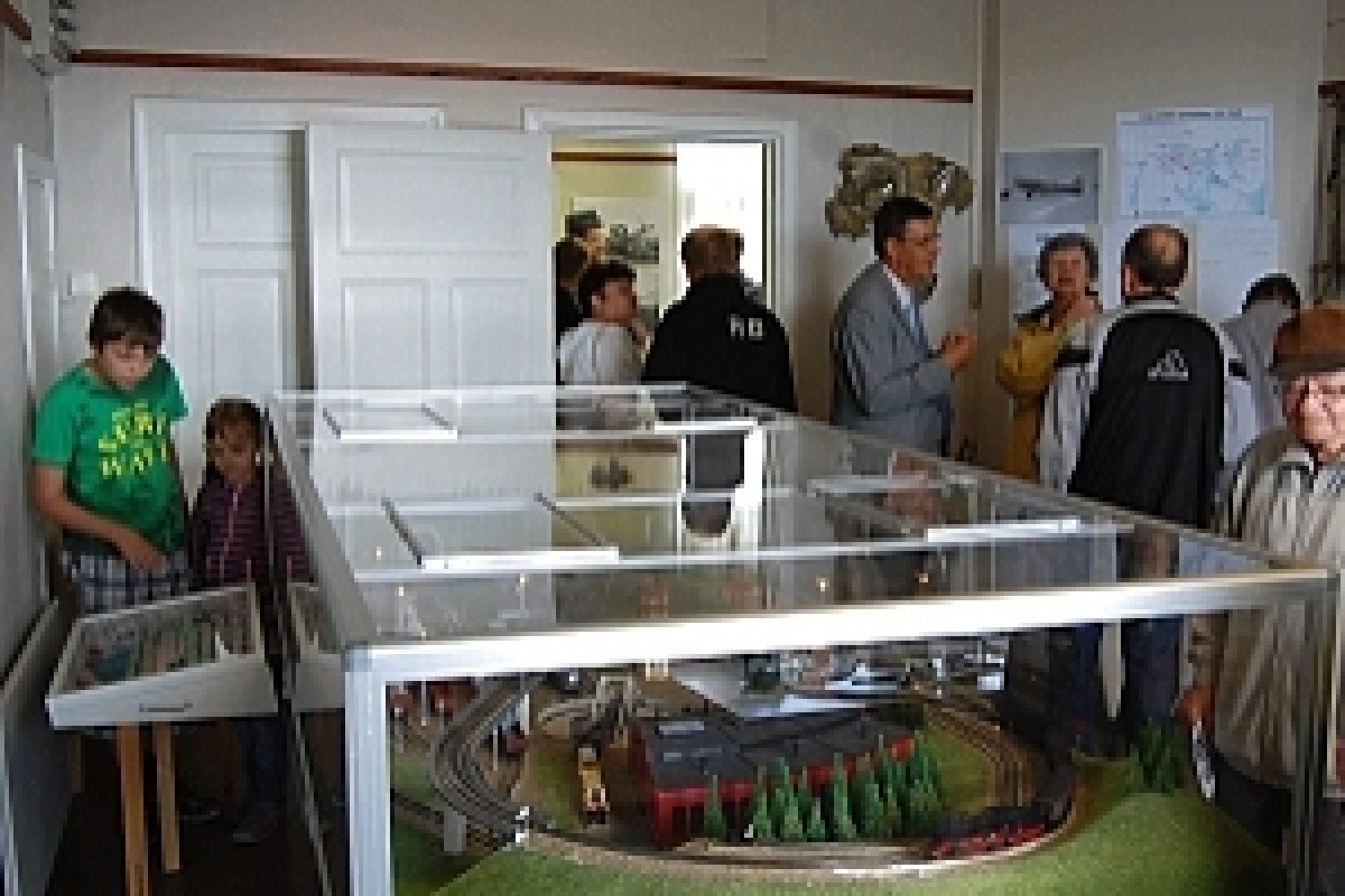 Nuoria ja vanhoja ihmisiä näyttelytilassa, jossa on vitriinin sisässä pienoisrautatie.