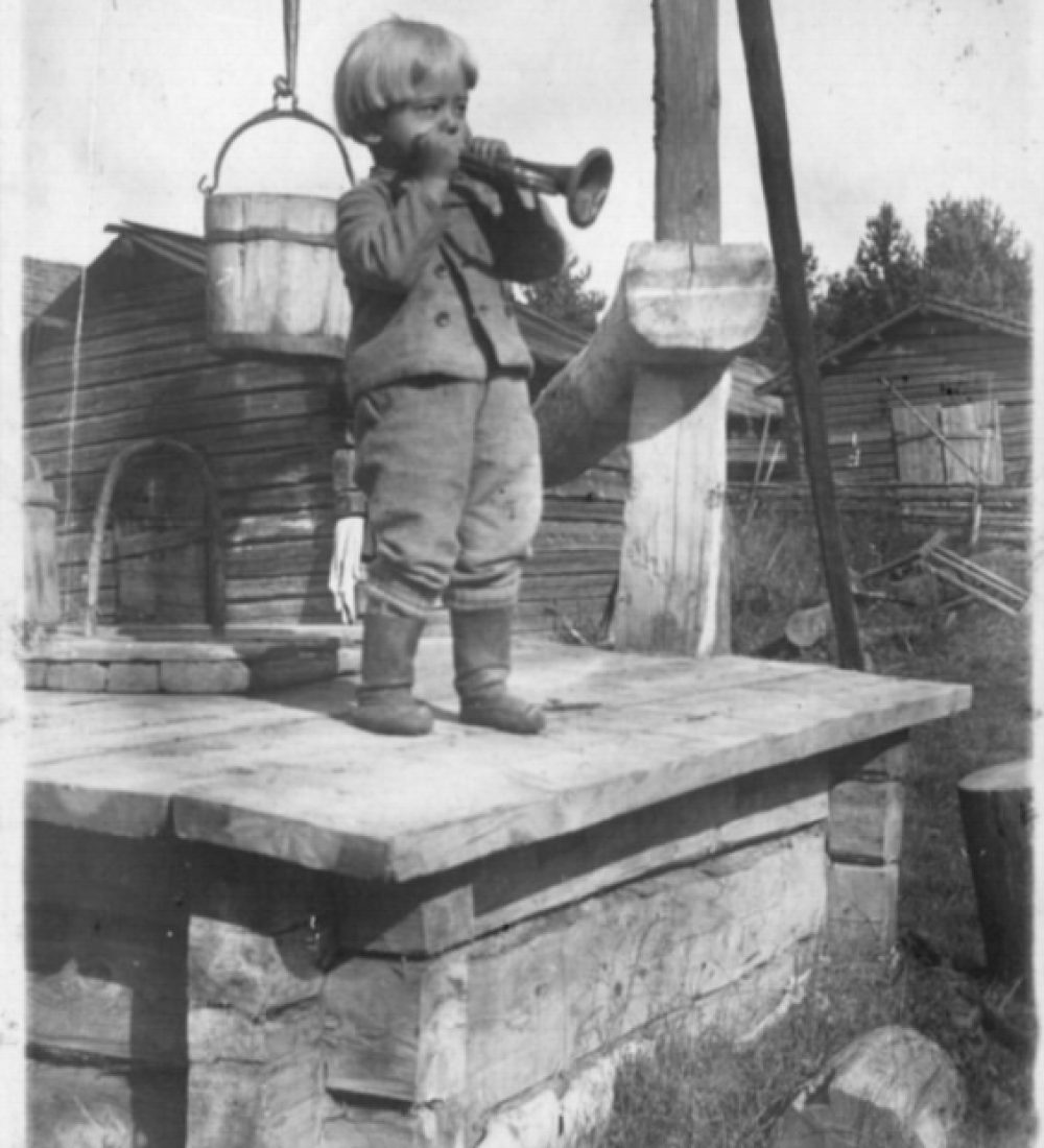 Pieni poika soittamassa torvea vinttikaivon kannella maalaistalon pihassa. Taustalla hirsisiä piharakennuksia. Savonlinnan maakuntamuseo.