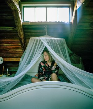 Nainen istuu puisessa, hunnulla verhotussa sängyssä ikkunan alla. Kuva näyttää olevan puutalon vintiltä tai muuten korkeimmasta kerroksesta.