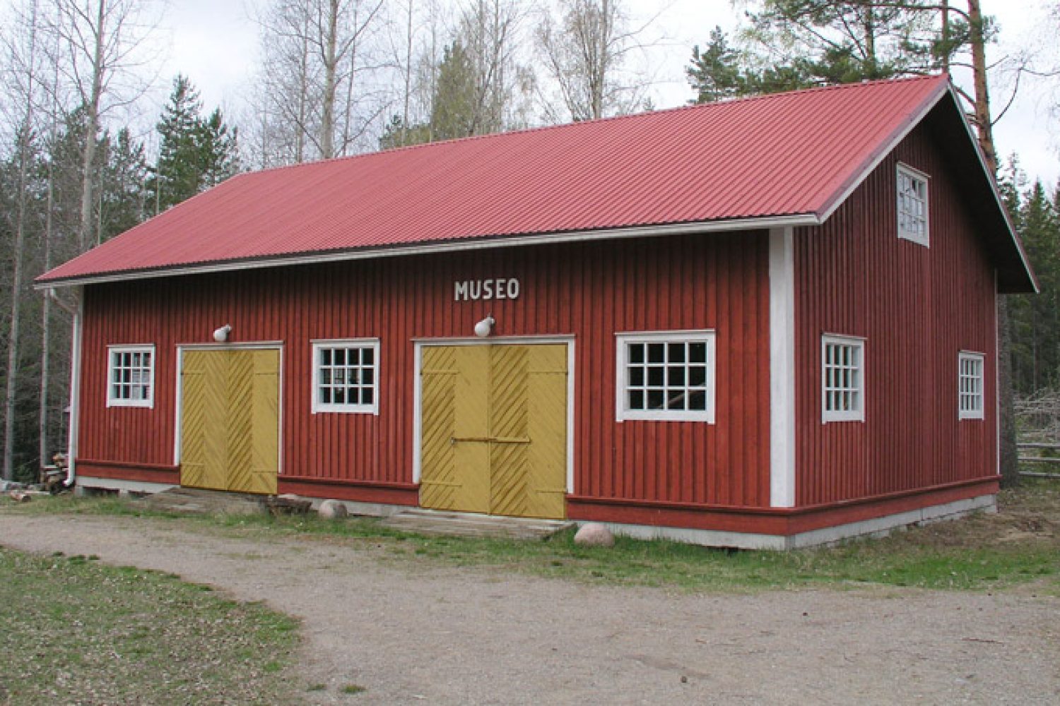 Punainen puutalo varustettuna peltikatolla ja kahdella keltaisella pariovella. Toisen oven yläpuolella on teksti "MUSEO".