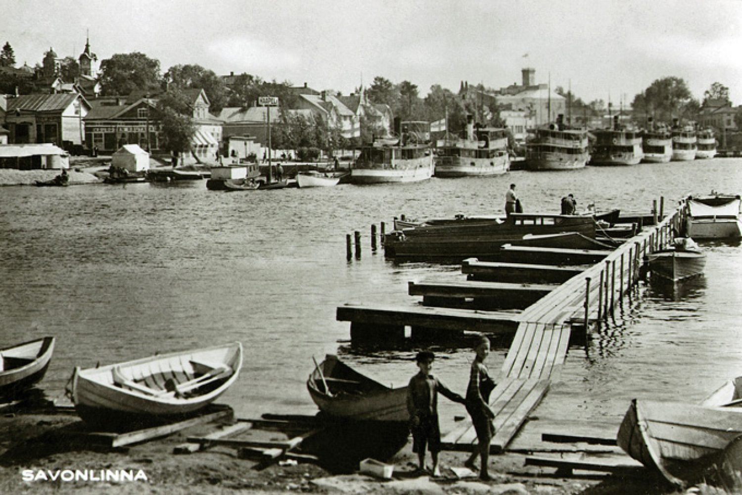 Mustavalkoisessa kuvassa näkyy kaupungin satamalaituri, jossa on seitsemän valkokylkistä höyrylaivaa ja pienempiä aluksia. Etualalla kuvassa on pienempi laituri, jossa on muutama soutuvene.