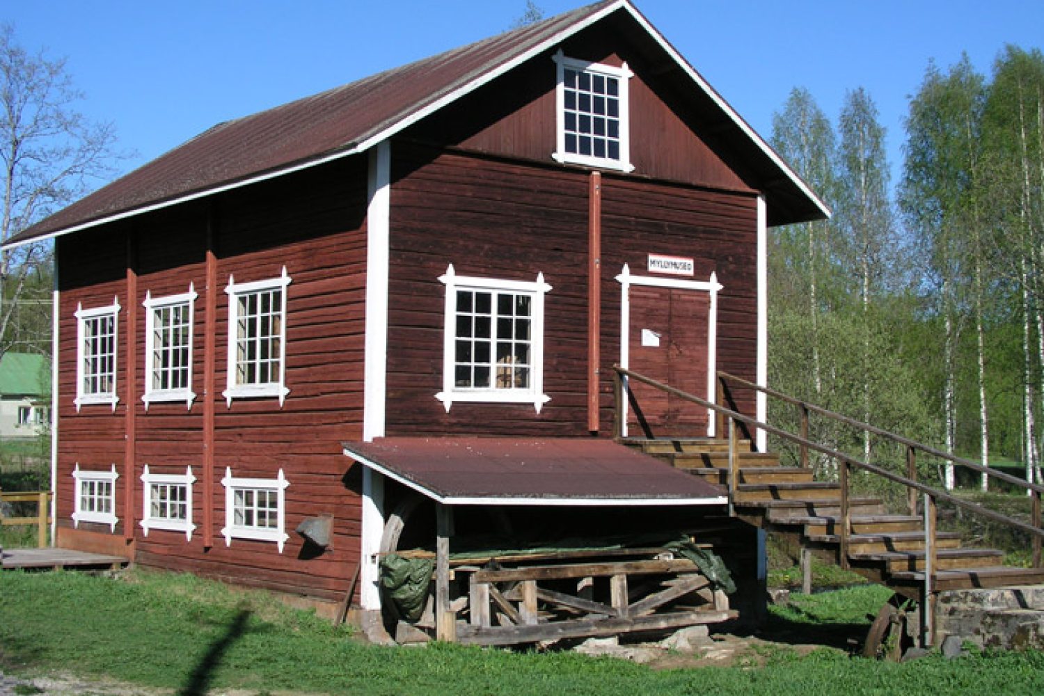 Punainen kaksikerroksinen hirsirakennus, jonka ovelle johtaa portaikko. Rakennuksessa on paljon ikkunoita.